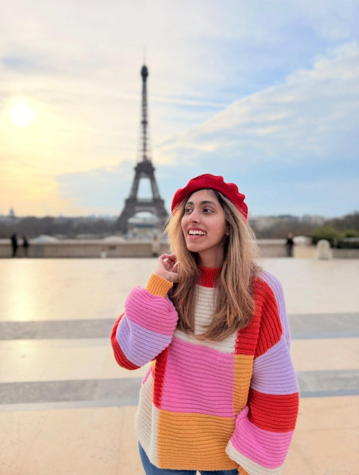 Woman in bright-colored sweater stands in front of the Eiffel Tower
